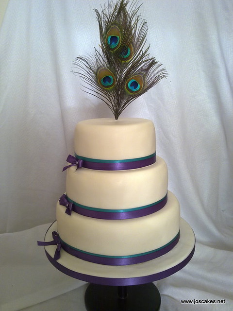 A three tier wedding cake with peacock feather topper trimmed with ribbons
