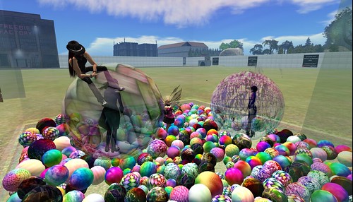 Several people stand inside of a nearly clear box filled with a wide variety of textured balls, most of them variations on rainbows. Two of the people are inside larger balls, and one of the three is sitting on top of one of the larger balls.