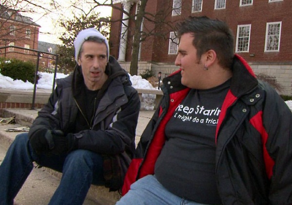 Dan Savage sits on steps outside of a college campus building with an overweight man in his early twenties. There is snow on the ground and they are both wearing winter clothes.