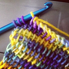 Close-up of the scarf in progress - showing one corner being worked off a blue metal crochet hook.