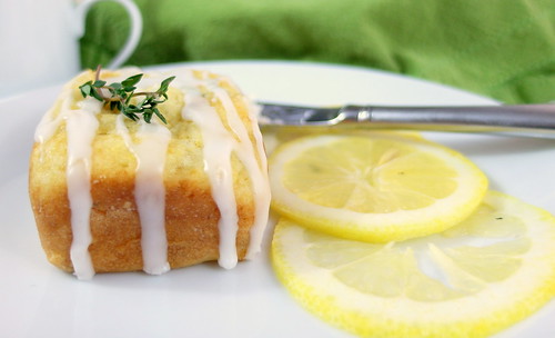 A glazed Lemon Thyme Tea Cake on a white plate with thin slices of lemon next to it. Garnished with a sprig of thyme.
