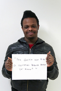 A black man stands with a sign that reads "you don't act like a normal black person, ya know?"