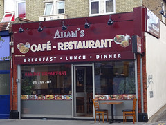 A small end-of-terrace shopfront with a large burgundy sign reading “Adam’s Café – Restaurant / Breakfast · Lunch · Dinner”.  Below is a glazed frontage with a slightly off-centre door in the middle.  A small table with two chairs is outside on the pavement.