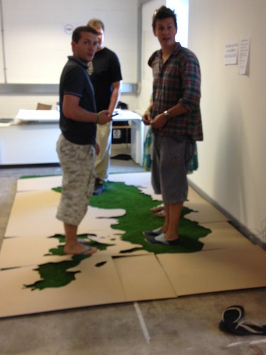 Three men stand on an astroturf and cardboard map.