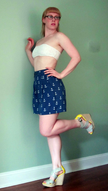 a blonde girl with glasses wearing a white bandeau top, blue anchor print shorts, and a gray and yellow wedges poses pinup style against a mint green wall