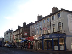 A terrace of buildings of different heights and styles, with various shopfronts along the ground floor, viewed obliquely on a late afternoon in winter.