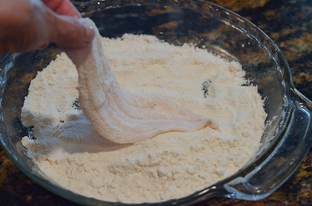 A hand dips raw fish into flour.