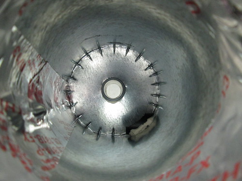 Looking up into the cyclone. The ring of spikes is the series of self-drilling sheet-metal screws holding the lid on, and is deliberately above the incoming airflow level. The black tube is the intake from the Protomat, and the grey tube is the exhaust to the vacuum.