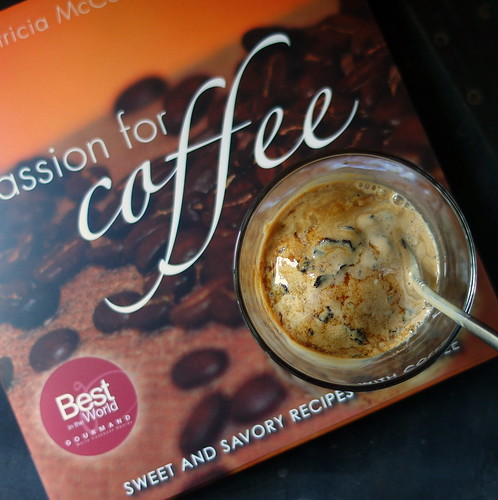 Overhead view and close up of front cover of Cook book: Passion for Coffee by Patricia McCausland-Gallo with a serving of Cafe Bomba in a clear glass with a long metal spoon. 