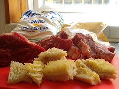 Three types of meat set out on a red chopping board: a piece of beef, some small bone-in lamb chops, and some chopped honeycomb tripe.  In the background is a bag of white flour with “1.5 kgs pounded iyan” printed on it and another bag containing a coarse brown powder.