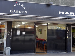 A shopfront with black boards almost to the very top.  A door in the middle is open, showing tables and chairs inside.  Old signage above reads “hisha Garden” (the “S” is missing).