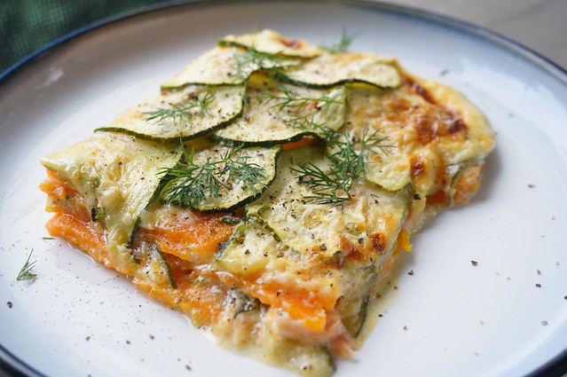 A close-up of a slice of scalloped sweet potatoes and zucchini with salmon. On the cut edge, the orange and green layers look like geological strata, and bits of fresh dill scattered on top look like tiny trees.