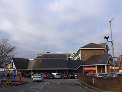 A view across a car park to a large but fairly low building with notably peaked roofs.  “Lidl” and “easyGym” branded signs are visible, and an office block with similar roofs rises behind.