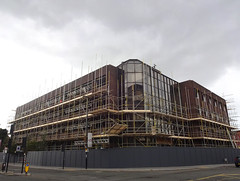 A large building at the junction of two streets.  The corner of the building is fully glazed, while the frontages stretching away down the two streets are dark brown brick or cladding.  The entire building is covered with scaffolding, and the ground floor is thoroughly boarded off from the pavement.
