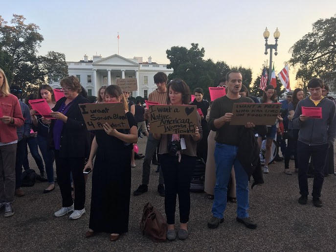 A group of people stand holding pink signs in front of the White House. In the foreground, there are two women and a man with cardboard signs.