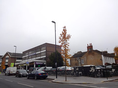 An oblique view onto three sets of buildings alongside a road.  To the left is a portion of a fairly modern-looking building with a pointed roof.  In the centre is a four-storey building with a very plain, rectangular profile, and plentiful windows on the frontage but none on the visible side wall.  The foreground to the right of this has no buildings, and instead several cars parked inside a low wall.  Behind the cars are terraced houses stretching back away from the main road.