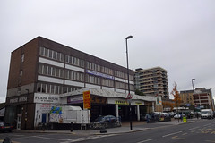 A wide-angle view of several large buildings along one side of a street.  In the foreground is a four-storey building with “Praise House” signs on it, and a protruding canopy on the ground floor with advertisements for car wash and tyre businesses.  Other, taller buildings are visible in the distance.
