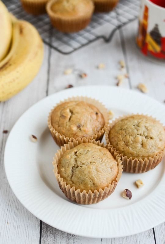 3 banana nut muffins on a white plate; coffee mug, bunch of bananas, and more muffins on a cooling rack in the background
